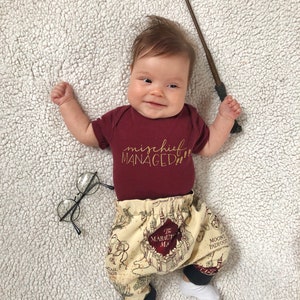 May include: A baby wearing a maroon onesie with the text "mischief managed" in gold lettering. The baby is wearing brown and beige pants with a map print of Hogwarts. The baby is holding a brown wand in one hand and wearing round black glasses.