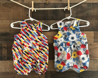 May include: Two baby overalls hanging on a clothesline. One has a geometric pattern in red, yellow, blue, and black. The other has a floral pattern in red, yellow, blue, and white.