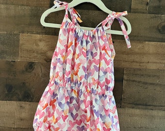 May include: A white sleeveless baby onesie with a pink, purple, and orange watercolor heart print. The onesie has a gathered elastic waistband and tie straps.