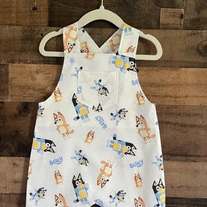 May include: White overalls with a blue and orange cartoon dog print. The overalls have a single pocket on the front and adjustable straps.