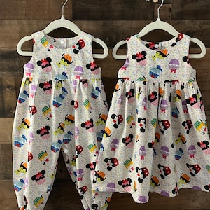 May include: Two white sleeveless dresses with a colorful print of cupcakes and Mickey Mouse heads. One dress is a jumper with a snap closure and the other is a dress with a gathered skirt.
