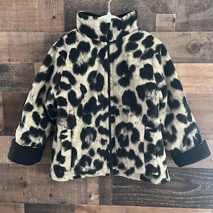 May include: A children's fleece jacket with a leopard print pattern in black and tan. The jacket has a high collar, a full-length zipper, and black cuffs. It is hanging on a white hanger against a wooden background.