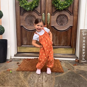 May include: A young child wearing an orange jumpsuit with a white top and pink shoes stands on a brown doormat. The jumpsuit has a repeating pattern. The background includes a wooden door with green wreaths and a sign that says "be THANKFUL".