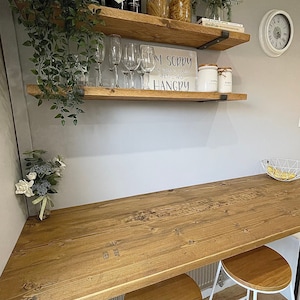 May include: Two wooden shelves with metal brackets mounted on a light gray wall. The shelves are decorated with plants, glasses, bottles of liquor, jars of pasta, and a sign that reads "I'm sorry for what I said when I was hangry".