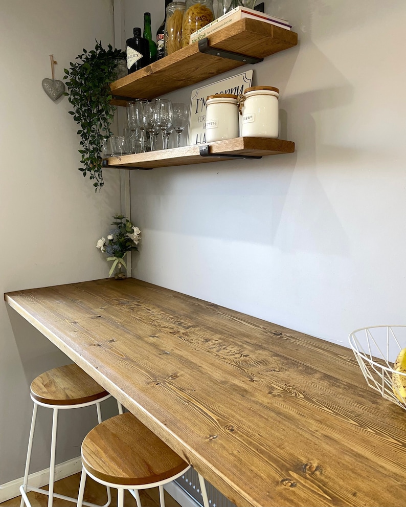 May include: Two white bar stools with wooden seats sit in front of a long wooden countertop. Two shelves with metal brackets are mounted on the wall behind the countertop. The shelves are filled with various items, including glasses, jars, and bottles. The text "I'm sorry for what I said when I was hungry" is printed on a sign on the top shelf.