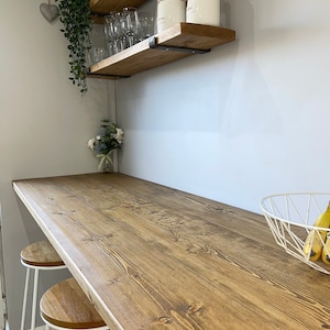 May include: A wooden countertop with a natural wood finish, two white bar stools, and a white wire basket with bananas. The countertop is attached to a wall with two wooden shelves, one with glasses and the other with jars labeled "Coffee" and "Sugar".