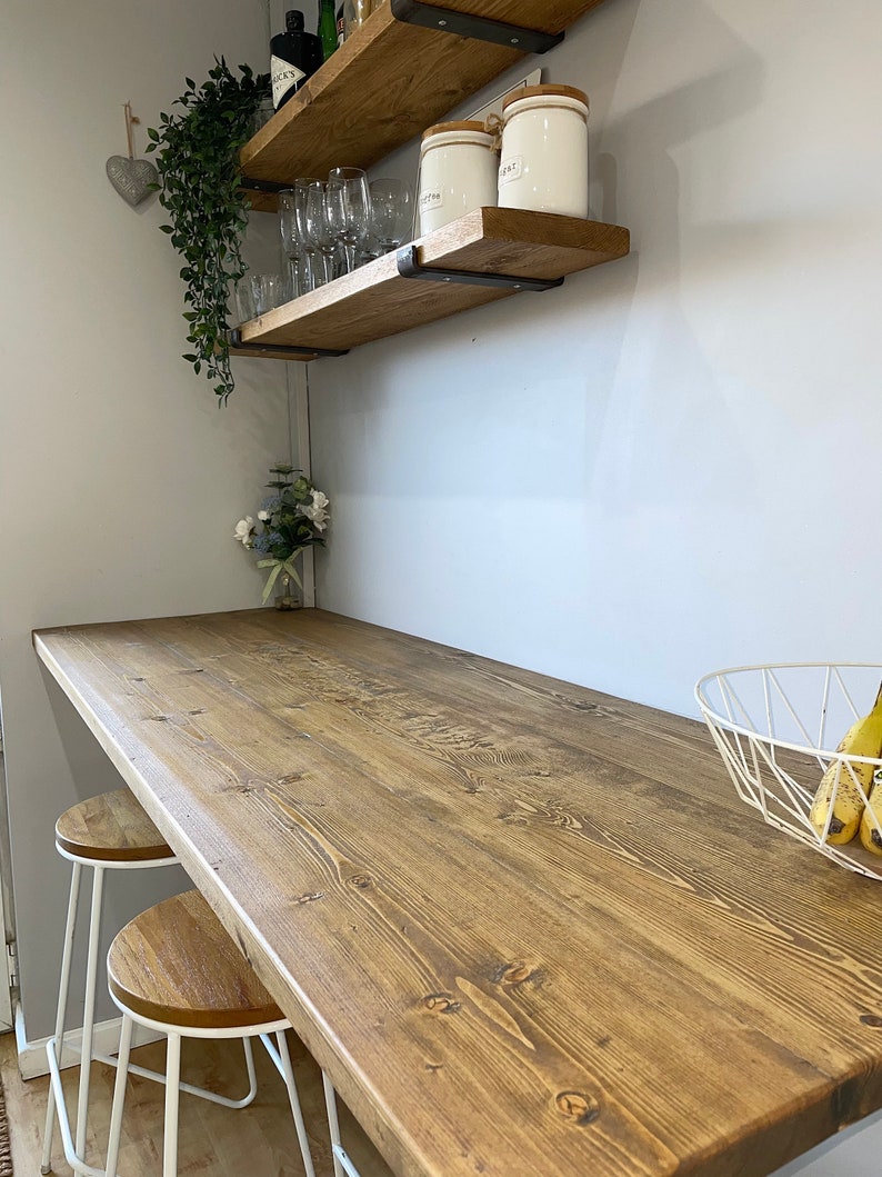 May include: A wooden countertop with a natural wood finish, two white bar stools, and a white wire basket with bananas. The countertop is attached to a wall with two wooden shelves, one with glasses and the other with jars labeled "Coffee" and "Sugar".