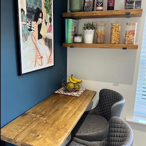 May include: A wooden floating shelf with black metal brackets, holding various items, including jars of pasta, a white planter with a lace design, and a cookbook. The shelf is mounted on a white wall, next to a blue wall with a framed print of a woman sitting at a cafe. A wooden table with a tiled surface is in the foreground, with two gray upholstered chairs. A bowl of fruit sits on the table.