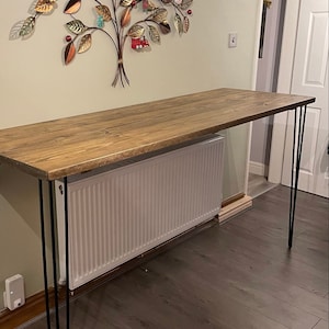 May include: A rustic wooden table with a natural wood finish and black hairpin legs. The table is placed in front of a white radiator and a wall decorated with a metal tree sculpture.