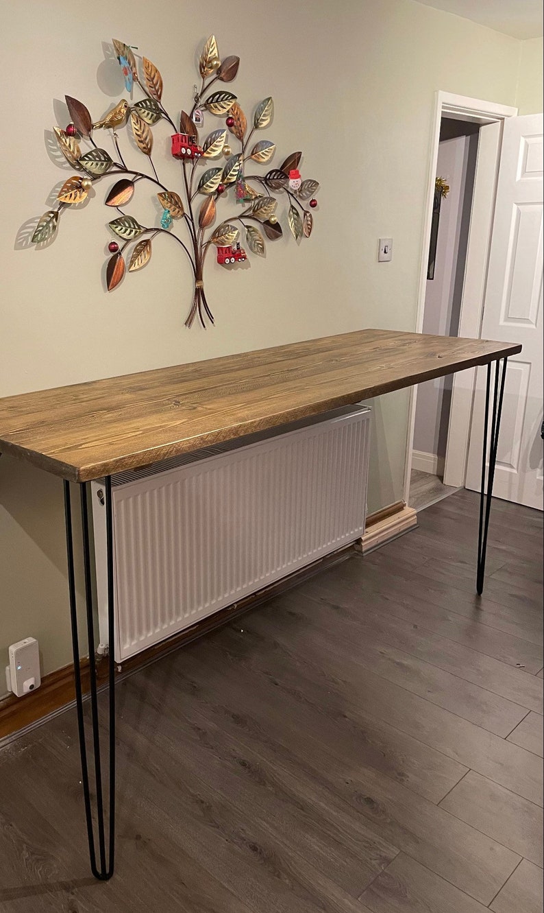 May include: A rustic wooden table with a natural wood finish and black hairpin legs. The table is placed in front of a white radiator and a wall decorated with a metal tree sculpture.