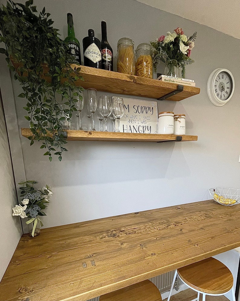 May include: Two wooden shelves with metal brackets mounted on a light gray wall. The shelves are decorated with plants, glasses, bottles of liquor, jars of pasta, and a sign that reads "I'm sorry for what I said when I was hangry".