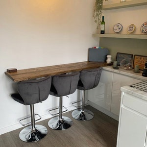 May include: Three gray velvet bar stools with chrome bases and footrests are positioned in front of a wooden countertop. The countertop is attached to a white wall and extends into a corner. The countertop is in a kitchen with white cabinets and a marble countertop.
