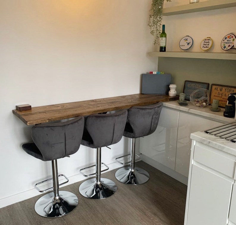 May include: Three gray velvet bar stools with chrome bases and footrests are positioned in front of a wooden countertop. The countertop is attached to a white wall and extends into a corner. The countertop is in a kitchen with white cabinets and a marble countertop.