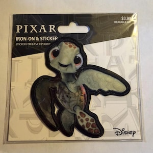 May include: A Pixar iron-on and sticker featuring a cartoon sea turtle character with a friendly expression. The sticker is in a clear plastic package with the Disney logo and the words "Wearable Art".