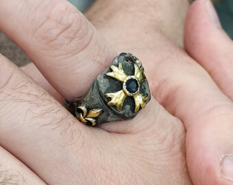 Puede incluir: Un anillo de plata con un diseño de cruz de oro y una piedra preciosa azul en el centro. El anillo se lleva en un dedo.