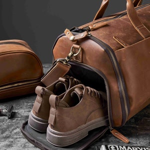May include: A brown leather duffel bag with a pair of brown shoes inside. A matching toiletry bag is to the left. A tube of toothpaste and a razor are also visible.