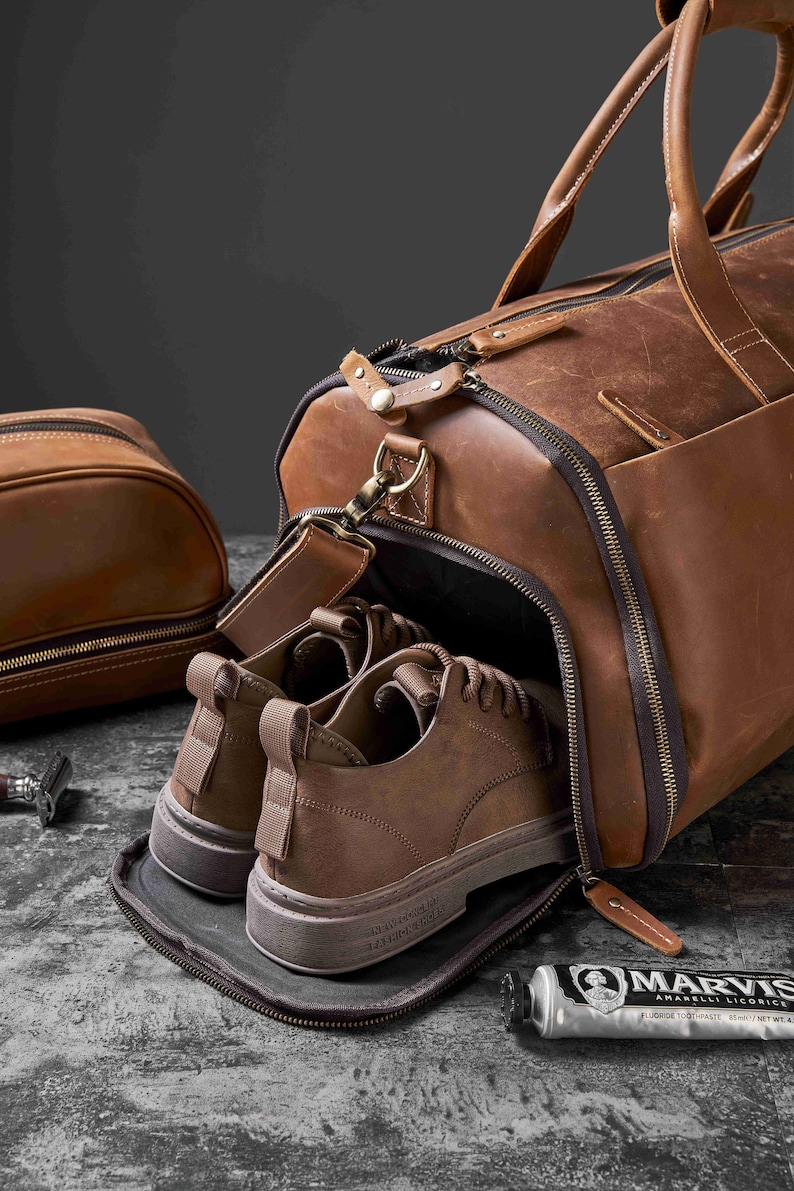 May include: A brown leather duffel bag with a pair of brown shoes inside. A matching toiletry bag is to the left. A tube of toothpaste and a razor are also visible.