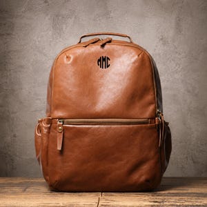 Puede incluir: Una mochila de cuero color canela con un asa superior, dos correas para los hombros y un bolsillo frontal con cremallera. La parte superior de la bolsa tiene grabadas las letras negras del monograma MMC. El diseño incluye bolsillos laterales y cremalleras de tono latón.