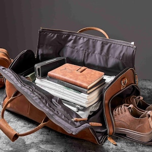 May include: Open brown leather travel bag with a variety of items inside, including a speaker, books, and a pair of brown shoes. A smaller brown leather bag is visible in the background.
