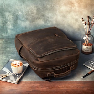 Pode incluir: Uma mochila de couro marrom com um bolso frontal com z&iacute;per e uma al&ccedil;a superior. A bolsa est&aacute; em uma mesa ao lado de uma x&iacute;cara de caf&eacute; e um caderno. O couro tem uma apar&ecirc;ncia desgastada.