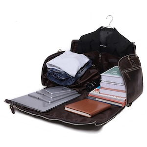 May include: Open brown leather travel bag with a black blazer on a hanger, folded clothes, a laptop, tablets, a smartphone, books, and a brown notebook. The bag is unzipped, revealing the contents.
