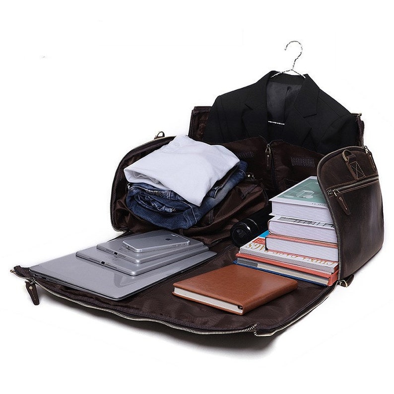 May include: Open brown leather travel bag with a black blazer on a hanger, folded clothes, a laptop, tablets, a smartphone, books, and a brown notebook. The bag is unzipped, revealing the contents.