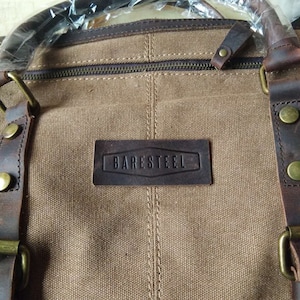 May include: A brown canvas travel bag with leather accents and a brown leather patch that says "BARESTEEL".