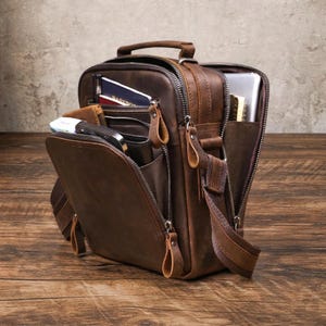 Könnte beinhalten: Eine braune Leder-Umhängetasche mit mehreren Fächern, Reißverschlüssen und einem Schultergurt. Die Tasche ist geöffnet und zeigt eine Brieftasche, einen Reisepass und andere Gegenstände. Die Tasche hat einen Griff oben und steht auf einer Holzoberfläche.