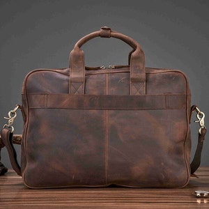 May include: A brown leather briefcase with a top handle and a detachable shoulder strap. The bag has a rectangular shape with a front pocket and metal hardware. The leather has a distressed finish.