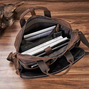 May include: Open brown leather briefcase with multiple compartments, including a padded laptop sleeve. The bag has a shoulder strap and top handles. Inside are books, a tablet, and other items. The bag is on a wooden surface.