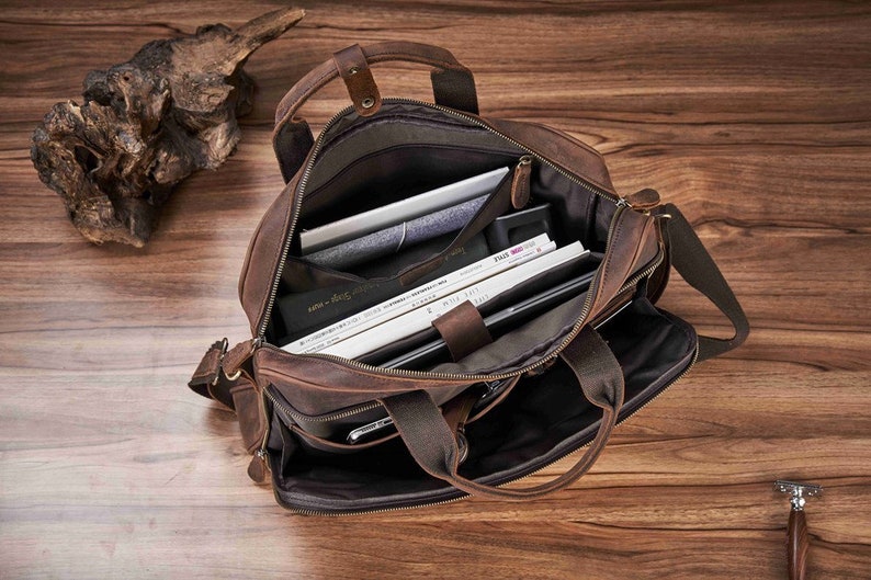 May include: Open brown leather briefcase with multiple compartments, including a padded laptop sleeve. The bag has a shoulder strap and top handles. Inside are books, a tablet, and other items. The bag is on a wooden surface.