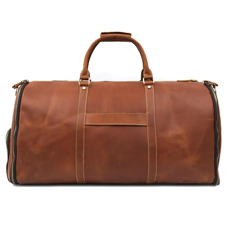 May include: A brown leather duffel bag with a top handle and two side zippers. The bag has a rectangular shape with a small rectangular patch on the front. The bag is made of a smooth, textured leather.