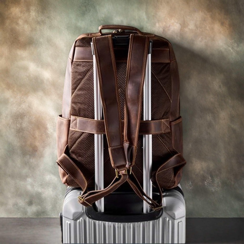 Pode incluir: Uma mochila de couro marrom com al&ccedil;as ajust&aacute;veis e uma al&ccedil;a superior, apoiada em uma mala de rodinhas prateada. A mochila tem um painel traseiro de malha e v&aacute;rios bolsos. Bagagem de viagem.