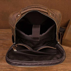 Pode incluir: Vista aberta de uma mochila de couro castanho com uma pega superior e v&aacute;rios compartimentos. O interior &eacute; castanho escuro com uma manga acolchoada para port&aacute;til. O exterior tem fecho de correr e um acabamento envelhecido.