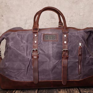 May include: A large, gray duffel bag with brown leather accents. The bag features brown leather handles, straps, and a bottom panel. The initials "RTB" are embossed on a leather patch. A zippered pocket is visible on the side.