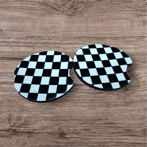 May include: Two car coasters with a black and white checkered pattern. The coasters are oval-shaped with a small notch on one side. The background is a wooden surface.