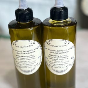 May include: Two amber glass bottles with black caps and white labels. The labels have the text "Creationscometrue" and list ingredients for a hair restoration oil blend. The ingredients include rosemary, fenugreek, alma, castor oil, sunflower oil, sweet almond oil, olive oil, tea tree oil, peppermint oil, and vitamin E oil.