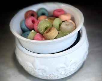 Könnte beinhalten: Weißer Keramik-Wachsschmelzer mit dekorativem Design. Die obere Schale ist mit bunten Wachsschmelzen gefüllt, die wie Donut-Ringe geformt sind.