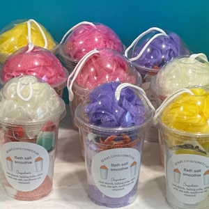 May include: Eight clear plastic cups filled with colorful bath salts and bath poufs. Each cup has a label that reads "Bath salt smoothie" and lists the ingredients.