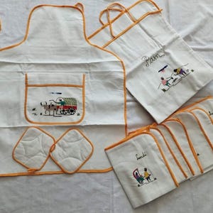Könnte beinhalten: Ein Set weißer Küchenwäsche mit orangefarbenem Besatz. Das Set beinhaltet eine Schürze mit einer Tasche, zwei Topflappen und acht Geschirrtücher. Die Schürze hat ein gesticktes Design mit einem von Ochsen gezogenen Wagen. Die Geschirrtücher haben gestickte Designs mit Menschen und Tieren. Das Set ist mit "Pam" und "Familia" beschriftet.