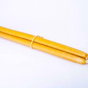 May include: Two long, slender, yellow candles tied together with a thin, light-colored string. The candles have a smooth surface and white wicks. The background is a plain white.