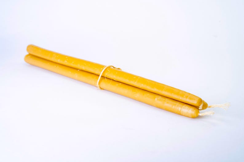 May include: Two long, slender, yellow candles tied together with a thin, light-colored string. The candles have a smooth surface and white wicks. The background is a plain white.