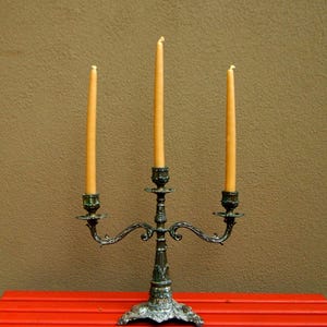 May include: An antique candelabra with three lit, slender, yellow candles. The candelabra is made of ornate, dark metal with a decorative base and curved arms. The candles are set against a brown background and a red table.
