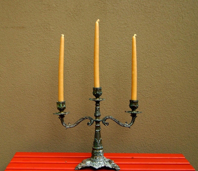 May include: An antique candelabra with three lit, slender, yellow candles. The candelabra is made of ornate, dark metal with a decorative base and curved arms. The candles are set against a brown background and a red table.
