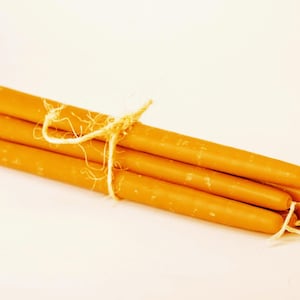 May include: A bundle of four orange-yellow taper candles tied together with natural twine. The candles have a smooth, cylindrical shape and are presented against a soft, off-white background. Ideal for home decor or special occasions.