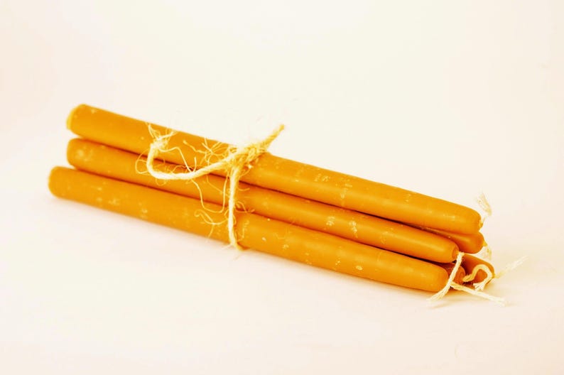 May include: A bundle of four orange-yellow taper candles tied together with natural twine. The candles have a smooth, cylindrical shape and are presented against a soft, off-white background. Ideal for home decor or special occasions.