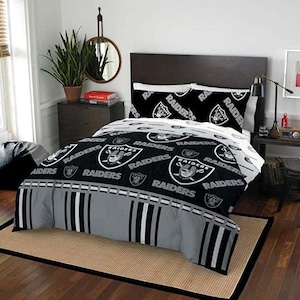 May include: A black, white, and gray bedding set featuring the Las Vegas Raiders logo and the word Raiders in a repeating pattern. The comforter includes a wide gray band at the foot with vertical black and white stripes, paired with two matching pillowcases.