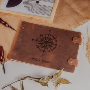 Op de afbeelding: Een bruin lederen gebonden dagboek met een kompasontwerp en de tekst "Captain Sparrow" gegraveerd op de voorkant.