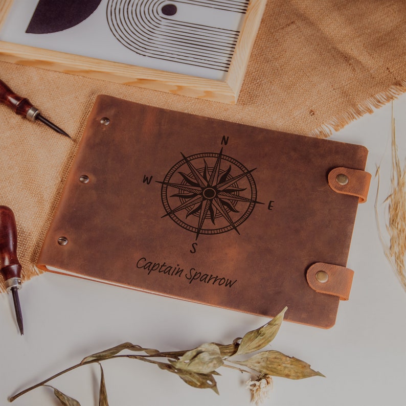 Op de afbeelding: Een bruin lederen gebonden dagboek met een kompasontwerp en de tekst "Captain Sparrow" gegraveerd op de voorkant.