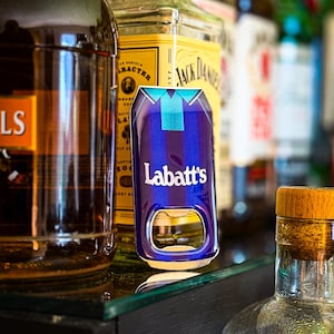 Könnte beinhalten: Ein blau-silberner Labatt's Flaschenöffner mit einem Ausschnitt zum Öffnen von Flaschen. Der Öffner befindet sich vor mehreren Spirituosenflaschen, darunter Jack Daniel's.